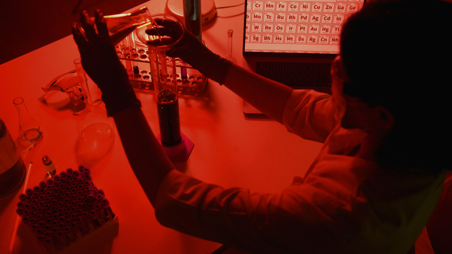 Chemist pouring a solution into a test tube in a lab lit in red, periodic table visible on a screen